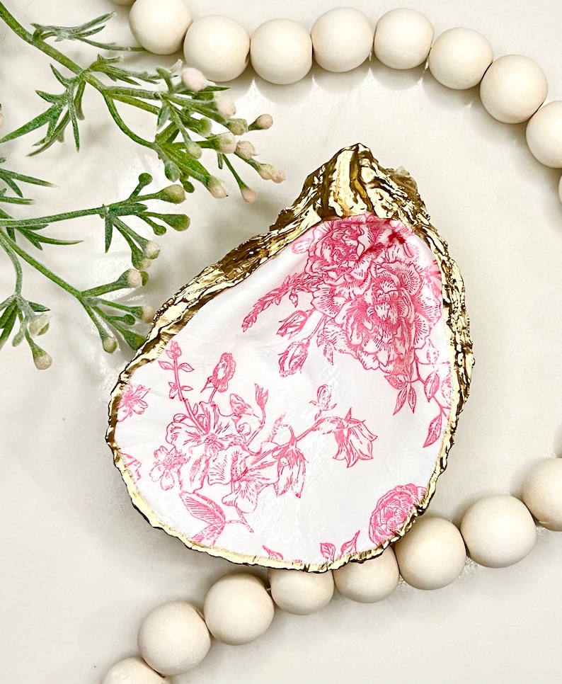 May include: A gold-rimmed oyster shell dish with a pink floral pattern. The dish is sitting on a white surface with a string of wooden beads around it.
