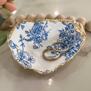 May include: A white ceramic ring dish with a blue floral pattern and a gold rim. Two gold rings are sitting in the dish.