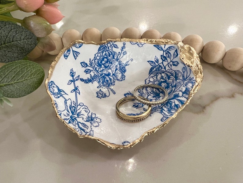 May include: A white ceramic ring dish with a blue floral pattern and a gold rim. Two gold rings are sitting in the dish.