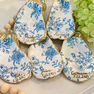 May include: Five white oyster shells with gold trim and blue floral patterns. Each shell has a gold inscription with a name and date, such as "Cassidy 7.12.2025".