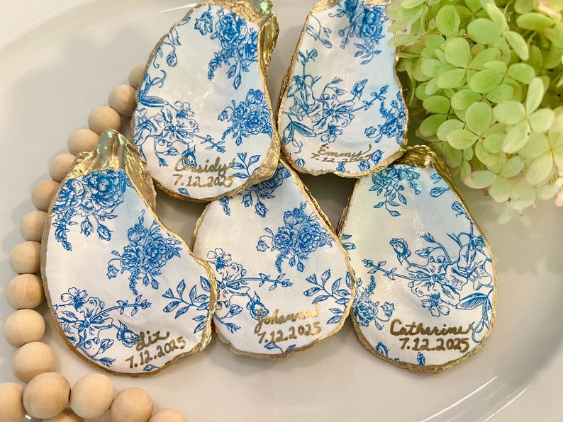 May include: Five white oyster shells with gold trim and blue floral patterns. Each shell has a gold inscription with a name and date, such as "Cassidy 7.12.2025".