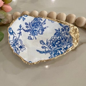 May include: A white ceramic oyster shell dish with a blue floral pattern and a gold rim. The dish is perfect for holding jewelry, keys, or other small items.