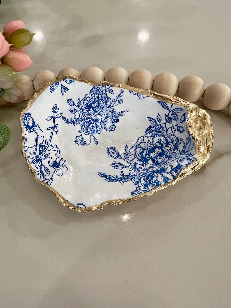May include: A white ceramic oyster shell dish with a blue floral pattern and a gold rim. The dish is perfect for holding jewelry, keys, or other small items.