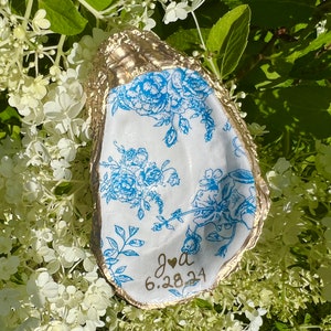 May include: A white oyster shell with a gold rim and blue floral pattern. The shell has the text "J & A 6.28.24" written on it.