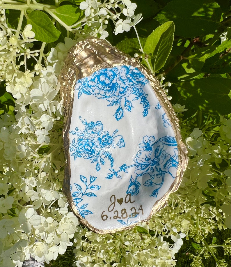 May include: A white oyster shell with a gold rim and blue floral pattern. The shell has the text "J & A 6.28.24" written on it.