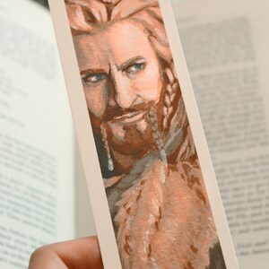 Bookmark print of younger dwarf brother painted in gouache. Bookmark is being held by hand over a background of an open fantasy book, a white blanket, plant, and small decorative photos.