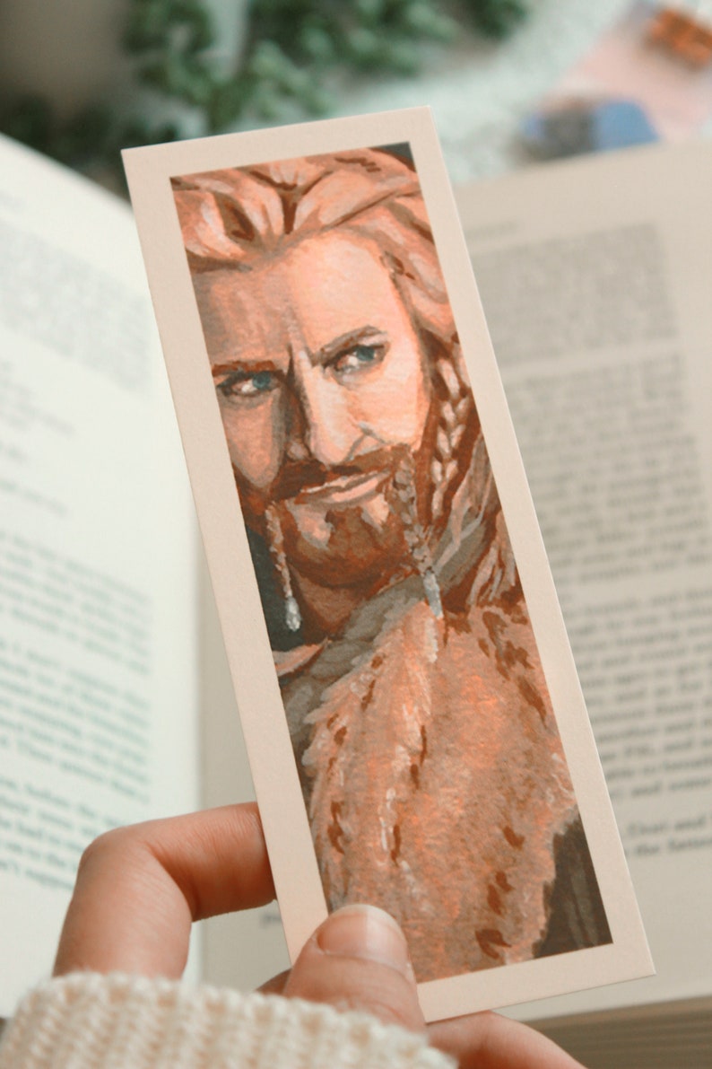 Bookmark print of younger dwarf brother painted in gouache. Bookmark is being held by hand over a background of an open fantasy book, a white blanket, plant, and small decorative photos.