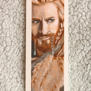 Bookmark print of older dwarf brother painted in gouache. Bookmark is on top of a plain background of a white blanket.