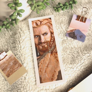 Bookmark print of younger dwarf brother painted in gouache. Bookmark is on top of a white fluffy blanket with a plant, small decorative photos, and the top of an open fantasy book in the background.