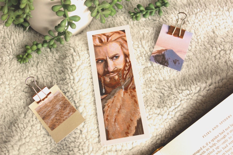 Bookmark print of younger dwarf brother painted in gouache. Bookmark is on top of a white fluffy blanket with a plant, small decorative photos, and the top of an open fantasy book in the background.