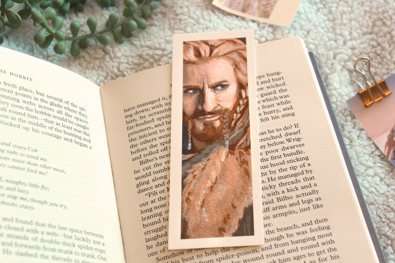 Bookmark print of older dwarf brother painted in gouache. Bookmark is on top of an open fantasy book with a white blanket, plant, and small decorative photos in the background.