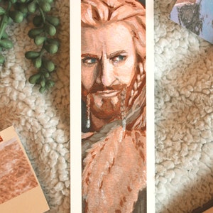 Bookmark print of older dwarf brother painted in gouache. Bookmark is on top of a white fluffy blanket with a plant, small decorative photos, and the top of an open fantasy book in the background.