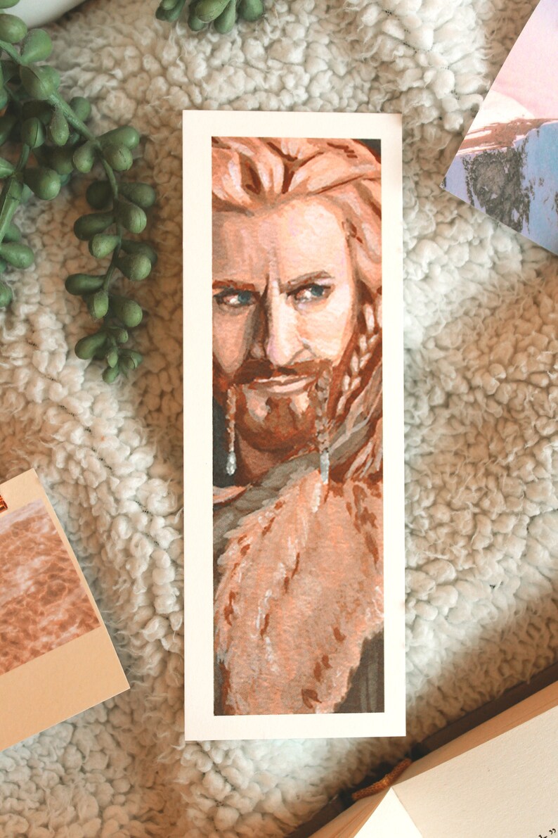 Bookmark print of older dwarf brother painted in gouache. Bookmark is on top of a white fluffy blanket with a plant, small decorative photos, and the top of an open fantasy book in the background.