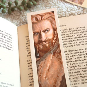 Bookmark print of older dwarf brother painted in gouache. Bookmark is on top of an open fantasy book with a white blanket, plant, and small decorative photos in the background.