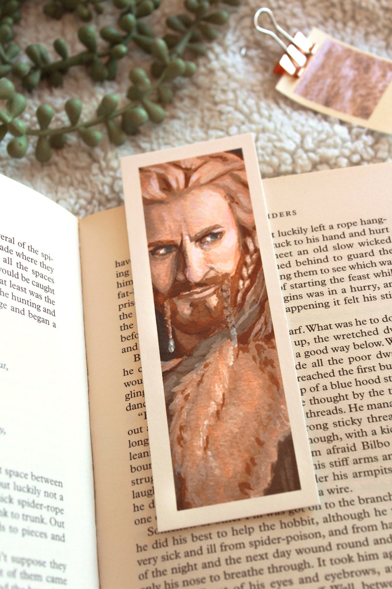 Bookmark print of older dwarf brother painted in gouache. Bookmark is on top of an open fantasy book with a white blanket, plant, and small decorative photos in the background.