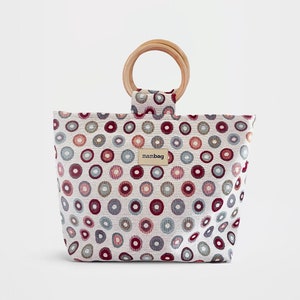 Puede incluir: Una bolsa de tela blanca con un patrón de círculos coloridos y un asa de madera. La bolsa tiene una pequeña etiqueta marrón con la palabra "mambag" en ella.