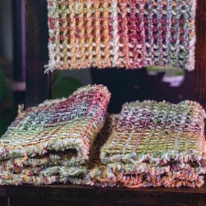 May include: Three rectangular woven fabric pieces featuring a variegated color palette of pink, green, yellow, and white. The textiles have a textured, open-weave grid pattern with visible yarn loops and frayed edges, displayed on a dark wooden surface.