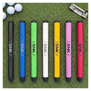 May include: Seven golf club grips in black, blue, green, white, yellow, pink, and red, each featuring a textured honeycomb pattern and the vertical text SNK with a stylized logo. The grips are arranged in a row alongside several white golf balls.