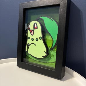 Può includere: Un'opera d'arte 3D incorniciata con un personaggio dei cartoni animati verde con una grande foglia e occhi rossi. L'opera d'arte è ambientata su uno sfondo verde pixelato e incorniciata in una scatola nera.