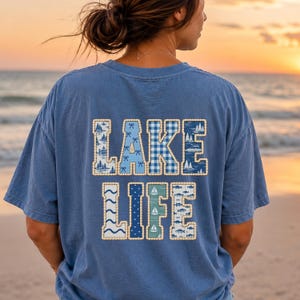 Puede incluir: Una camiseta azul de manga corta con el texto LAKE LIFE en la espalda. Las letras están rellenas con varios patrones en azul y blanco, incluyendo veleros, peces, lazos, cuadros y árboles, todo delineado con un borde punteado de color claro.