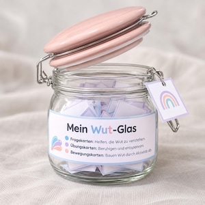 Op de afbeelding: Een doorzichtige glazen pot met een roze deksel en metalen sluiting, gevuld met kleine papieren kaartjes. Een label op de voorzijde toont de tekst Mein Wut-Glas met uitleg over kaarten voor woedebeheersing, ontspanning en beweging. Een vierkant label met een regenboog hangt aan de sluiting.