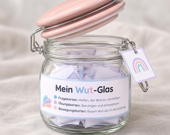 Puede incluir: Un frasco de vidrio transparente con tapa rosa y cierre metálico, lleno de pequeñas tarjetas de papel. Una etiqueta frontal dice Mein Wut-Glas con texto sobre tarjetas para entender la ira, relajarse y moverse. Una pequeña etiqueta cuadrada con un arcoíris cuelga del cierre.