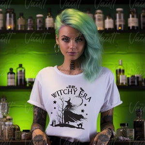 May include: White t-shirt with the text "IN MY WITCHY ERA" and a witch silhouette design. The design includes a witch, moon, stars, a raven, a cat, and a candle. The person in the image has green hair and tattoos.