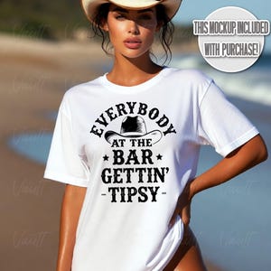 May include: White t-shirt with the text "EVERYBODY AT THE BAR GETTIN' -TIPSY-" in black. The design includes a cowboy hat graphic. The shirt is a short-sleeved crew neck.