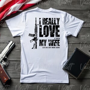 Peut inclure: T-shirt blanc avec un dessin noir d'un fusil et le texte "I REALLY LOVE IT WHEN MY WIFE LETS ME BUY MORE GUNS." Un pistolet et un livre sont &eacute;galement visibles.