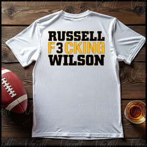May include: White t-shirt with black and yellow text that reads "Russell F3cking Wilson".