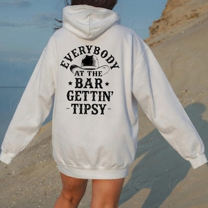 May include: A white hoodie with a black graphic on the back that reads "EVERYBODY AT THE BAR GETTIN' TIPSY" with a cowboy hat graphic. The hoodie has a hood and long sleeves.