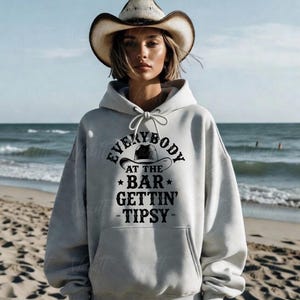 May include: A light gray hoodie with the text "EVERYBODY AT THE BAR GETTIN' TIPSY" in black. The hoodie features a drawstring hood and a kangaroo pocket. The person in the image is wearing a cowboy hat.
