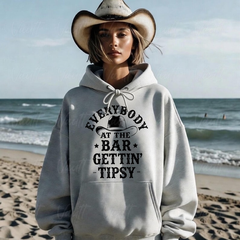 May include: A light gray hoodie with the text "EVERYBODY AT THE BAR GETTIN' TIPSY" in black. The hoodie features a drawstring hood and a kangaroo pocket. The person in the image is wearing a cowboy hat.