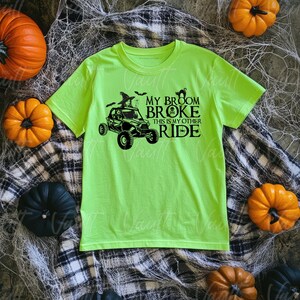 May include: A neon green t-shirt with a black graphic of a witch driving a side-by-side ATV. The text on the shirt reads "My Broom Broke, This is My Other Ride."