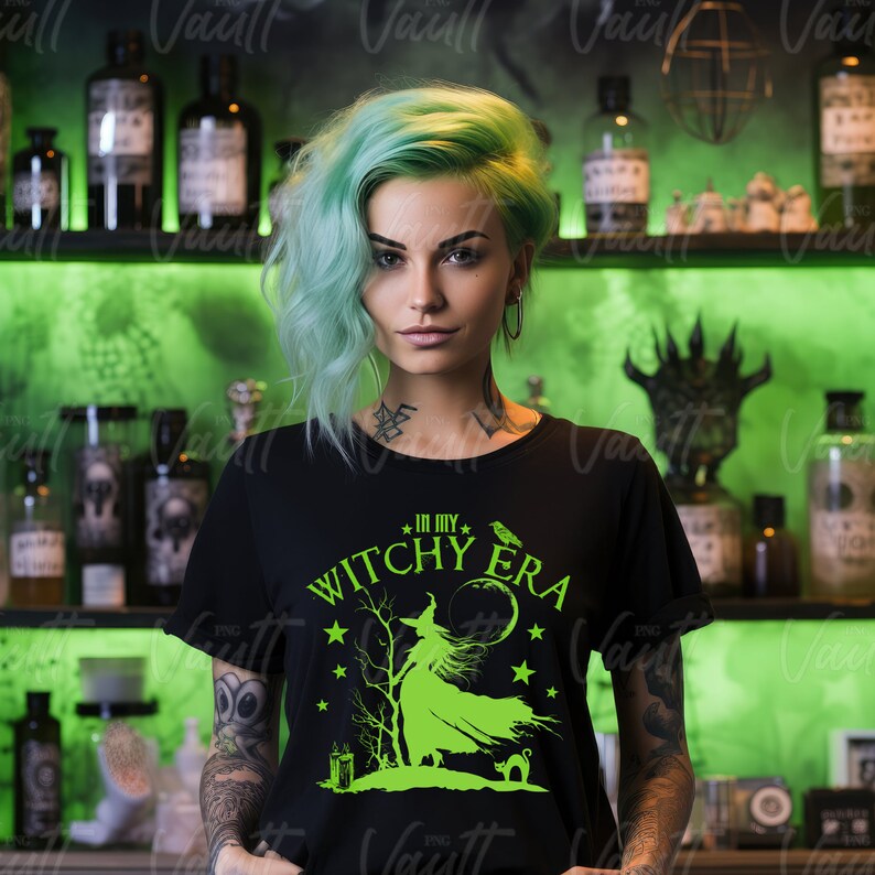 May include: Black t-shirt with the text "IN MY WITCHY ERA" in neon green. The graphic features a witch, a cat, a raven, stars, and a full moon. The woman in the image has green hair and tattoos.