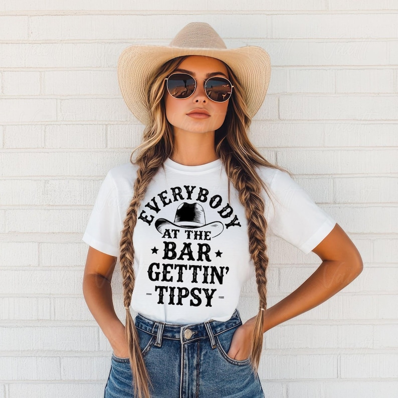 May include: White t-shirt with the text "EVERYBODY AT THE BAR GETTIN' TIPSY" in black. The shirt features a cowboy hat graphic. The person in the image is wearing a straw cowboy hat and sunglasses.