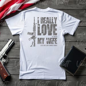 Peut inclure: T-shirt blanc avec un dessin gris d'un fusil et le texte : "I REALLY LOVE IT WHEN MY WIFE LETS ME BUY MORE GUNS." Un pistolet et un livre sont &eacute;galement visibles.