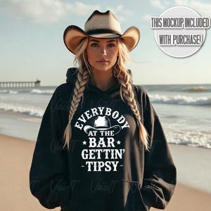 May include: Black hoodie with the text "EVERYBODY AT THE BAR GETTIN' -TIPSY-" in white. The design includes a cowboy hat graphic. The image also includes a cowboy hat.