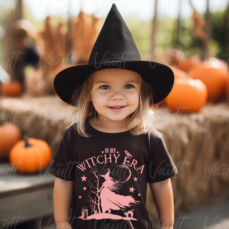 May include: A child wearing a black witch hat and a dark brown t-shirt with the text "IN MY WITCHY ERA" and a pink witch graphic. The background includes pumpkins and hay bales, creating a Halloween theme.