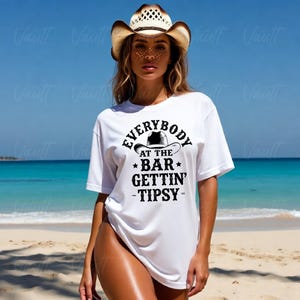 May include: White t-shirt with the text "EVERYBODY AT THE BAR GETTIN' -TIPSY-" in black. The shirt features a cowboy hat graphic. A straw cowboy hat is worn. The background shows a beach and ocean.