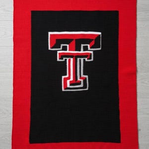 May include: A rectangular crochet blanket featuring a large black center panel with a red and white letter T logo. The blanket is framed by a wide, solid red border. The design is worked in a consistent, textured stitch pattern throughout the piece.