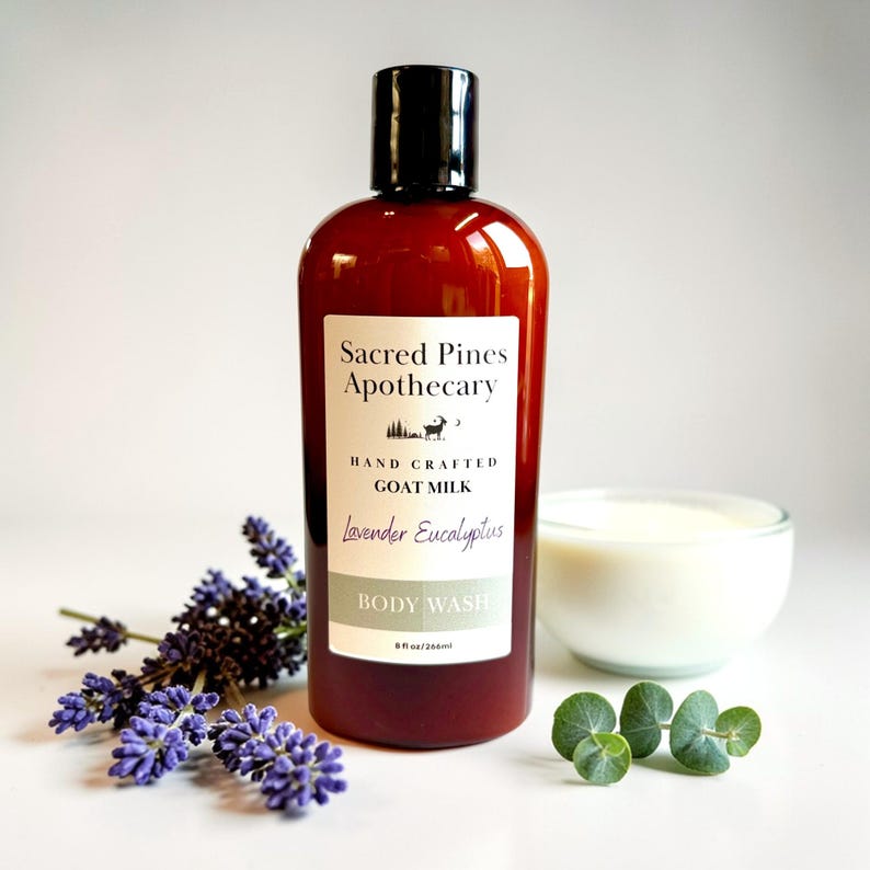 May include: A brown bottle of "Sacred Pines Apothecary" body wash with a black cap, 8 fl oz/236ml. The label reads "Hand Crafted Goat Milk Lavender Eucalyptus Body Wash." A small white bowl of cream, sprigs of eucalyptus, and lavender sprigs are also present.