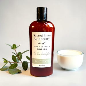 May include: A brown bottle of Sacred Pines Apothecary body wash with a black cap, labeled "Tea Tree Eucalyptus" and "Goat Milk." A small white bowl of cream and a sprig of green leaves are next to the bottle. The bottle contains 9 fl oz.