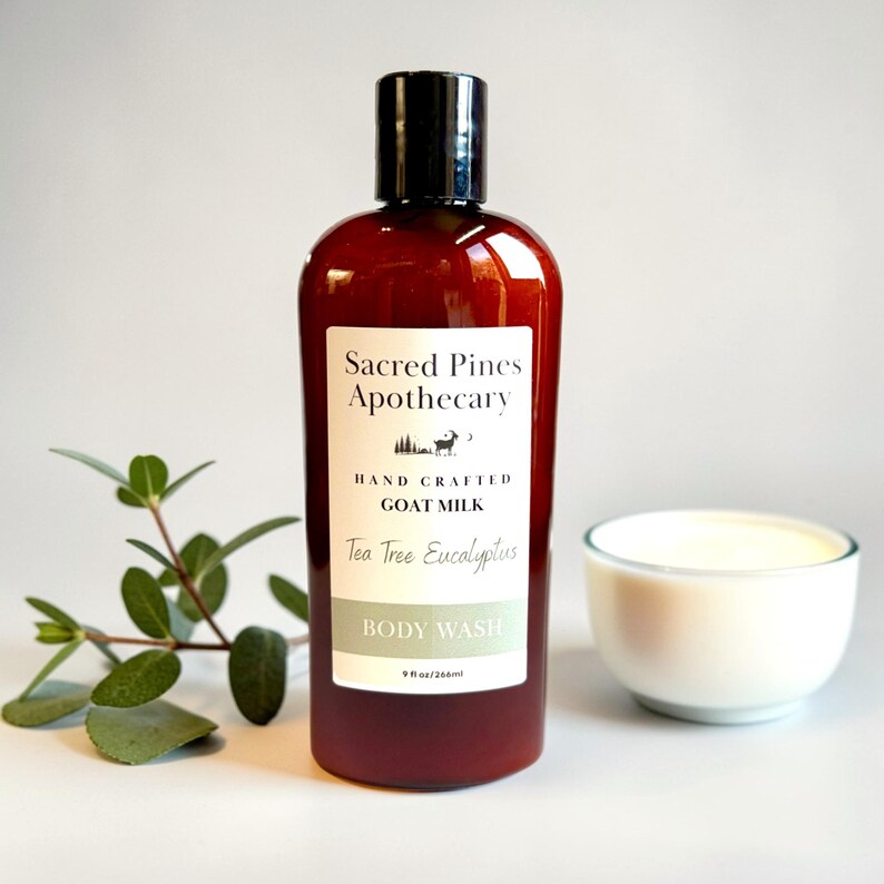 May include: A brown bottle of Sacred Pines Apothecary body wash with a black cap, labeled "Tea Tree Eucalyptus" and "Goat Milk." A small white bowl of cream and a sprig of green leaves are next to the bottle. The bottle contains 9 fl oz.
