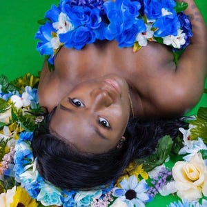 May include: A person surrounded by an arrangement of flowers, including blue, white, and yellow blooms, against a green backdrop. The person is adorned with a blue floral garland. The image has a soft, natural aesthetic.
