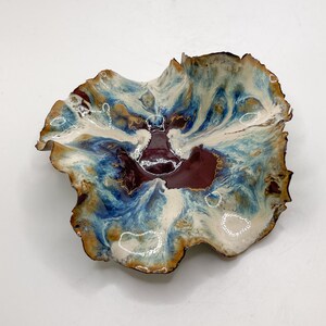 May include: A handmade ceramic bowl with an irregular, natural form. The bowl has a marbled pattern in blue, white, and burgundy. The rim is edged with dark brown. This decorative bowl is suitable for serving or display purposes.