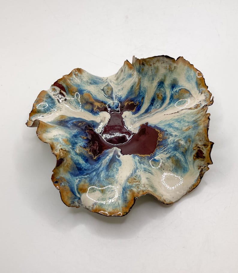 May include: A handmade ceramic bowl with an irregular, natural form. The bowl has a marbled pattern in blue, white, and burgundy. The rim is edged with dark brown. This decorative bowl is suitable for serving or display purposes.