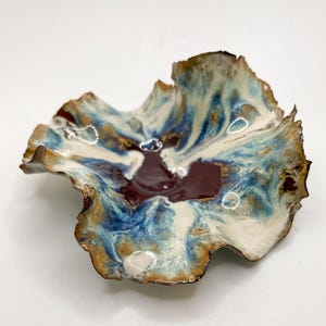 May include: A handcrafted ceramic bowl with an organic shape. The bowl has a rich colour palette of deep burgundy, swirling blues, creamy whites, and gold. The edges are gently ruffled, adding to its artistic appeal. This decorative bowl is ideal for serving or display.