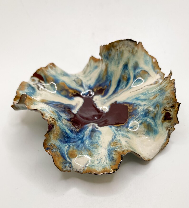 May include: A handcrafted ceramic bowl with an organic shape. The bowl has a rich colour palette of deep burgundy, swirling blues, creamy whites, and gold. The edges are gently ruffled, adding to its artistic appeal. This decorative bowl is ideal for serving or display.