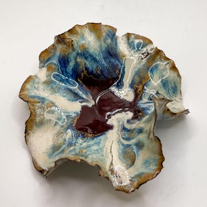 May include: A handcrafted ceramic bowl with an organic shape. The bowl has a mix of colours: deep burgundy, blues, cream, and brown. The edges are slightly raised, and the surface has a glossy finish. The bowl is approximately 25 cm in diameter.